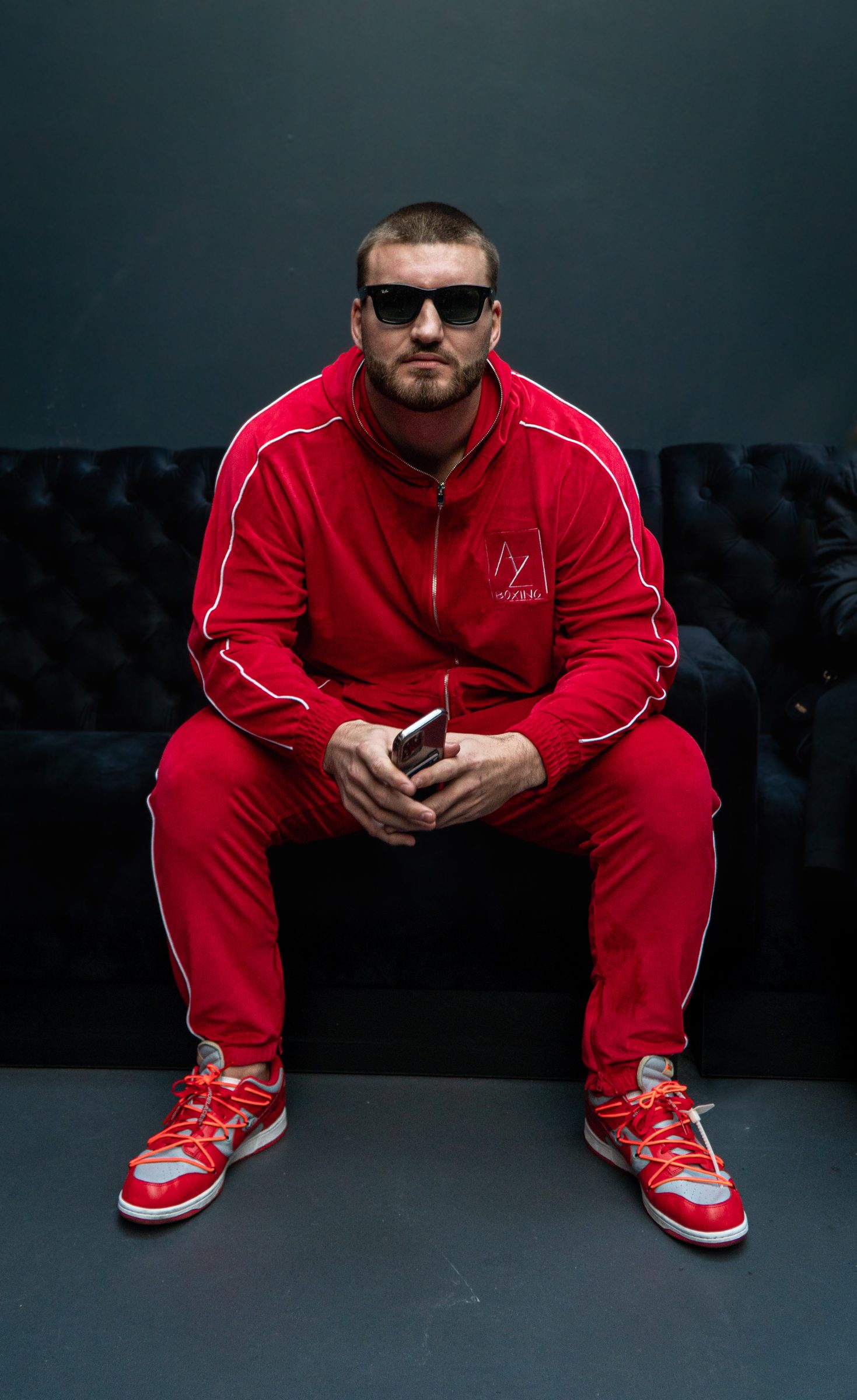 Fighter in a red tracksuit and sunglasses seated on a dark set