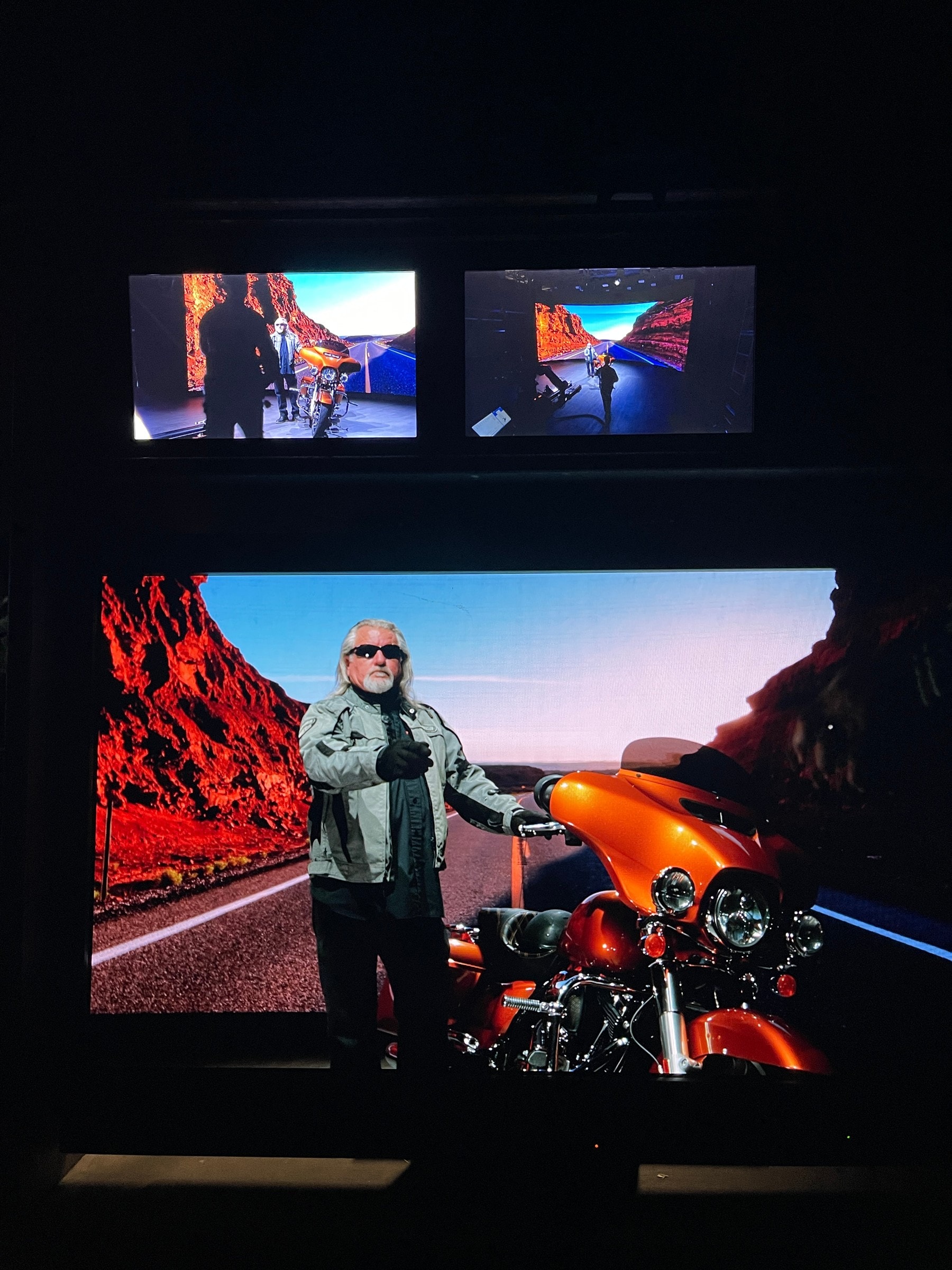 Rider beside an orange motorcycle on an LED desert road, with the camera feed on monitors above